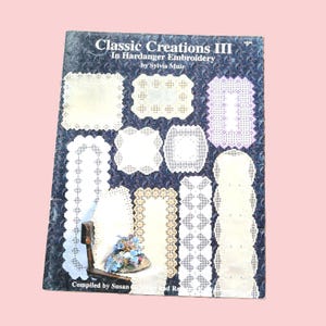 May include: A book titled "Classic Creations III In Hardanger Embroidery by Sylvia Muir" with various white and cream-colored embroidery patterns. The book cover is dark blue with a floral design. A small decorative chair with a floral arrangement is in the foreground.