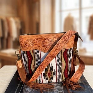 May include: A tan leather shoulder bag with a patterned fabric body. The bag features a decorative floral design on the leather, a long adjustable strap, and a fringe detail. The bag has a zip closure.