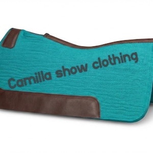 May include: A turquoise saddle pad with brown leather accents. The pad has the text "Camilla show clothing" printed on it in dark gray. The pad is designed for equestrian use.