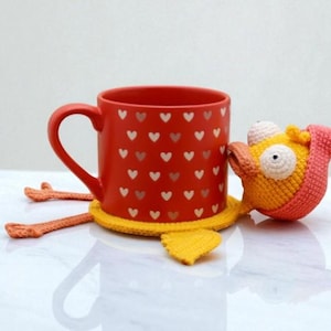 May include: A red ceramic mug decorated with white hearts rests on a yellow crocheted coaster, accompanied by a crocheted creature. The creature is yellow, orange, and pink, with large white eyes and a beak.
