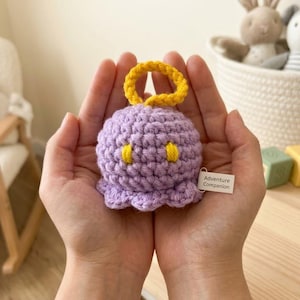 Puede incluir: Un juguete de crochet hecho a mano con forma de criatura morada con detalles amarillos y un halo amarillo. El juguete tiene una etiqueta que dice "Adventure Companion". El juguete se sostiene en manos abiertas.
