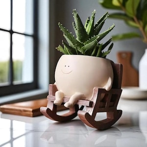 May include: A novelty beige plant pot in the shape of a character with a smiling face, seated in a brown rocking chair. The pot contains a green succulent plant. The design is decorative and suitable for indoor use.
