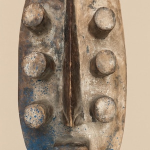 Puede incluir: Una máscara de madera ovalada y desgastada con una cresta vertical central y seis protuberancias cilíndricas. La máscara presenta una nariz y boca talladas, con pintura azul salpicando la superficie. El color general es una mezcla de marrón, beige y azul.