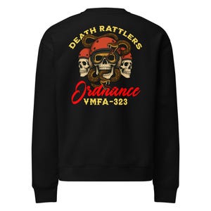 May include: Black sweatshirt with a graphic featuring three skulls wearing helmets, a snake, and the words "DEATH RATTLERS" and "Ordnance VMFA-323" in yellow and red. The design is centered on the back of the sweatshirt.