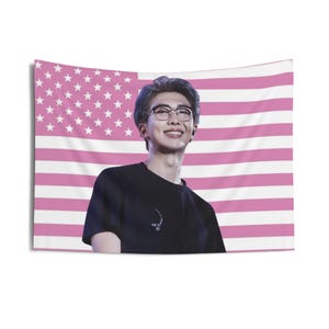 Puede incluir: Un tapiz rectangular con una persona sonriente con gafas y una camiseta negra, sobre un diseño de bandera estadounidense rosa y blanca. Adecuado para la decoración de paredes.
