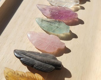 May include: A collection of carved gemstone feathers in various colors, including yellow, black, pink, green, and purple. The feathers are arranged on a light wooden tray, showcasing their intricate details and natural beauty. Ideal for home decor or as unique gifts.