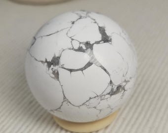 May include: A smooth, spherical white stone featuring a network of dark gray, web-like veins throughout its surface. The polished orb rests securely on a small, circular, light beige stand, showcasing its natural marbled appearance.