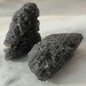 Puede incluir: Dos rocas de color gris oscuro, de textura rugosa y superficie ligeramente reflectante. Las rocas tienen formas irregulares y parecen formaciones naturales. La imagen es un primer plano que destaca los detalles de la superficie de la piedra.