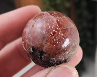 May include: A polished, spherical sunstone featuring a translucent, reddish-brown base with internal fractures and distinct, shimmering golden metallic inclusions scattered throughout the surface of the stone.