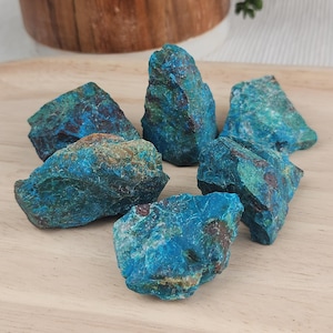 May include: A collection of six raw, unpolished chrysocolla mineral stones. Each stone features a vibrant mix of deep blue, turquoise, and earthy brown tones with a rough, jagged texture and irregular, angular shapes scattered across the surface.