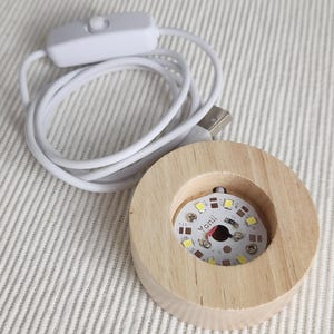 May include: A round light-colored wooden display base featuring a circular cutout in the center that houses a small white circuit board with multiple LED lights and the text Yonii. A white USB power cord with an inline switch is attached.