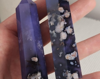 May include: Two polished crystal towers with a deep purple hue and translucent quality. The stones feature distinct white and light brown floral-like inclusions scattered throughout the smooth, faceted surfaces, creating a natural pattern.