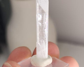 May include: A clear, elongated quartz crystal point standing vertically on a small, circular white clay base, which is attached to a transparent, hexagonal acrylic display stand. The crystal has a smooth, faceted surface.
