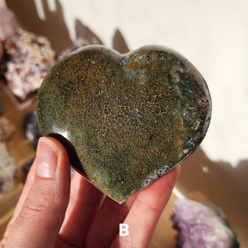 May include: A heart-shaped stone with a mottled pattern of green, brown, and white. The polished surface reflects light, highlighting the natural textures and colors of the stone. The letter "B" is visible at the bottom of the image.