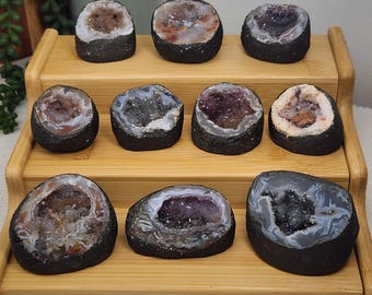 May include: A collection of nine geode halves with dark, rough outer shells and crystalline interiors in shades of purple, white, gray, and brown, arranged on a three-tiered wooden display stand with a smooth, light-toned finish.