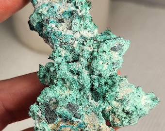 May include: A raw mineral specimen featuring a vibrant green, textured surface with crystalline formations and dark grey inclusions. The rock has an irregular, jagged shape with varying shades of emerald and mint green throughout its porous structure.