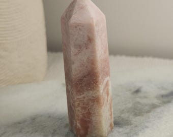 May include: A polished, six-sided crystal tower featuring a blend of soft pink, dusty rose, and creamy white mineral patterns. The stone has a smooth, opaque texture with a pointed, faceted top and a flat base, standing upright.