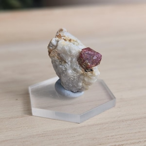 May include: A natural mineral specimen with a striking, deep red crystal set in a white and tan rock matrix. The mineral is displayed on a clear, hexagonal stand, highlighting its natural form and colour. The stand is clear.