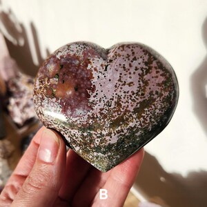 May include: A heart-shaped stone with a polished surface. The stone features a mix of colors, including shades of pink, brown, and green, with a speckled pattern. The letter "B" is visible on the bottom of the stone. The stone is held in a hand.