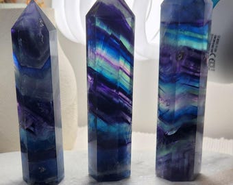 May include: Three polished fluorite crystal towers standing upright. Each hexagonal point features distinct, layered bands of deep purple, vibrant blue, and translucent green, showcasing natural color variations and internal mineral inclusions.