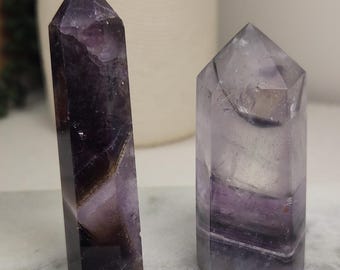 May include: Two polished amethyst crystal points with hexagonal bases. The taller point on the left displays deep purple and dark violet hues, while the shorter point on the right features lighter translucent lavender and clear quartz banding.