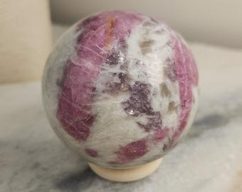 May include: A polished spherical stone featuring a mix of deep magenta and white mineral inclusions. The surface displays a smooth, rounded texture with visible crystalline patterns throughout the orb, resting on a small circular base.