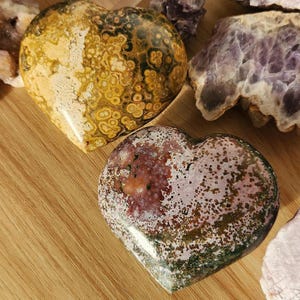 May include: Two heart-shaped stones with unique patterns. One is yellow and brown with a spotted design, the other is red, green, and white with a speckled appearance. The stones are displayed on a wooden surface, with other crystals in the background.