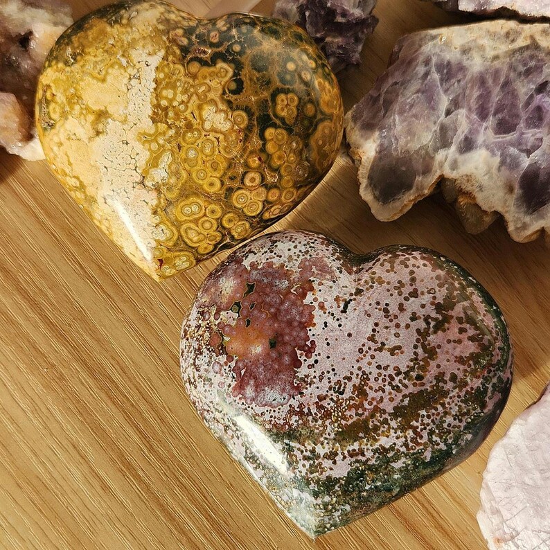 May include: Two heart-shaped stones with unique patterns. One is yellow and brown with a spotted design, the other is red, green, and white with a speckled appearance. The stones are displayed on a wooden surface, with other crystals in the background.