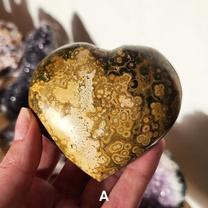 May include: A heart-shaped stone with a unique pattern of brown, tan, and dark green spots and swirls. The stone is held in a hand, and the letter "A" is visible at the bottom. The stone has a polished, smooth surface.