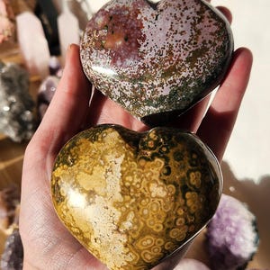 May include: Two heart-shaped stones are held in a hand. The top stone is a mix of burgundy, green, and tan. The bottom stone is a mix of tan, brown, and cream. Other crystals are visible in the background.