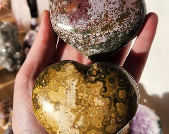 May include: Two heart-shaped stones are held in a hand. The top stone is a mix of burgundy, green, and tan. The bottom stone is a mix of tan, brown, and cream. Other crystals are visible in the background.