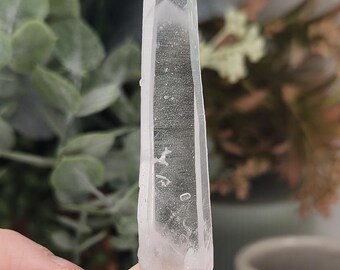 May include: A clear, elongated quartz crystal point with a hexagonal structure and a pointed termination. The crystal features natural internal inclusions, fractures, and a translucent appearance with varying degrees of clarity throughout the stone.