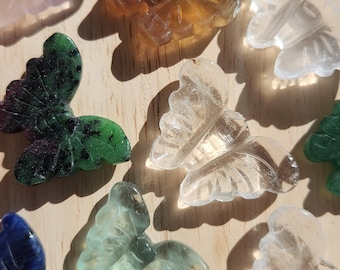 May include: A collection of carved butterfly figurines in various colors, including clear, pink, green, blue, and amber. The butterflies are displayed on a light-colored wooden surface, showcasing their intricate details.