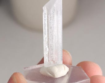 May include: A clear, elongated, prismatic crystal specimen standing vertically on a small, white, rounded clay base, which is attached to a transparent, hexagonal acrylic display stand for stability and viewing.