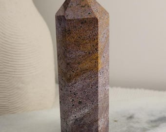 May include: A polished hexagonal crystal tower featuring a mottled pattern of earthy brown, deep purple, and muted yellow tones. The stone has a smooth, faceted surface with a pointed top, showcasing natural mineral inclusions and color variations.