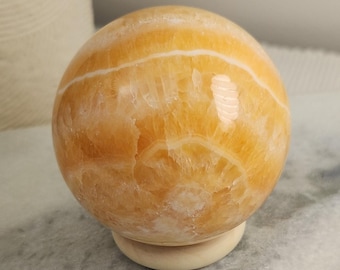 May include: A polished orange calcite sphere featuring natural white banding and crystalline textures, resting securely on a small, circular, light-colored wooden stand. The stone displays a smooth, glossy surface with translucent areas.