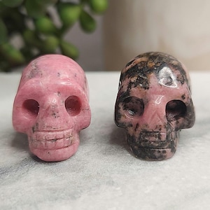 May include: Two carved gemstone skulls. One is a solid pink color, the other is pink with brown and black markings. The skulls have eye sockets, a nose cavity, and a detailed jawline. They are displayed on a light-colored surface.