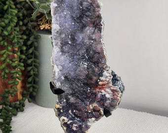 May include: A tall, vertical amethyst crystal cluster featuring deep purple and dark brown mineral formations. The specimen is mounted on a black metal stand with a square base and two support arms that hold the crystal upright for display.