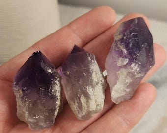 May include: Three raw amethyst crystal points with deep purple tips that fade into translucent, pale, and cloudy white bases. Each stone features natural jagged edges, crystalline textures, and pointed terminations, showcasing variations in color and clarity.
