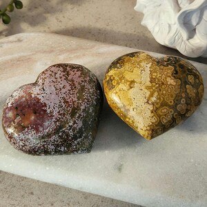 May include: Two heart-shaped stones. One is a mix of burgundy, green, and white, while the other is a blend of yellow, brown, and cream. The stones have a polished, smooth surface and are likely used for decorative or spiritual purposes.
