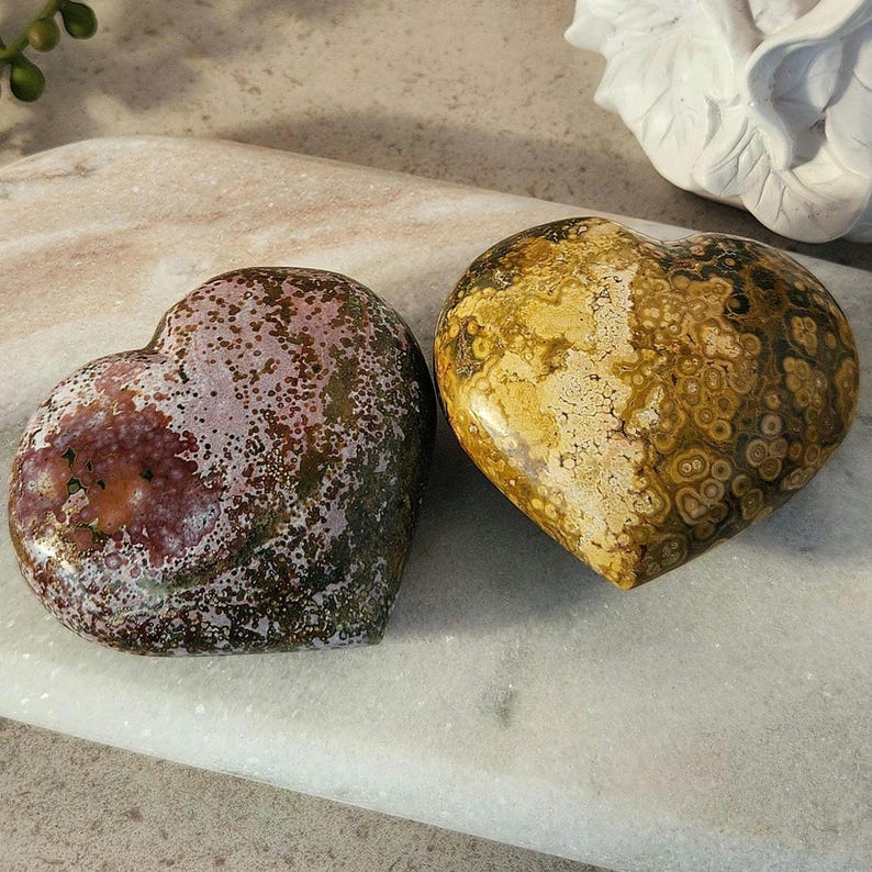 May include: Two heart-shaped stones. One is a mix of burgundy, green, and white, while the other is a blend of yellow, brown, and cream. The stones have a polished, smooth surface and are likely used for decorative or spiritual purposes.