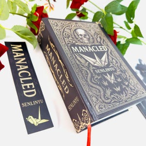 Op de afbeelding: Een donkergrijs boek met gouden letters en sierlijke ontwerpen, waaronder een schedel en origami-kranen. De titel "MANACLED" en auteur "SENLINYU" zijn zichtbaar. Een bijpassende bladwijzer met dezelfde tekst en een papieren kraan is ook aanwezig.