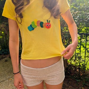 May include: A bright yellow short-sleeved t-shirt featuring a colourful felt caterpillar appliqué on the chest. The caterpillar is composed of several circular segments in shades of blue, green, yellow, orange, and red, with a red head and eyes.