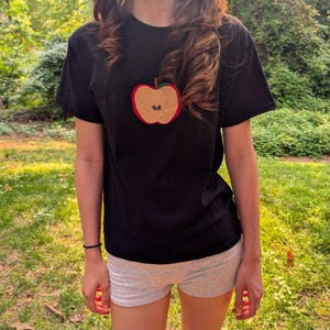 Może przedstawiać: Czarny t-shirt z krótkim rękawem z wyhaftowaną na środku grafiką jabłka. Wzór jabłka składa się z beżowego wnętrza z dwoma małymi czarnymi pestkami, czerwonej obwódki i brązowego ogonka. Koszulka posiada okrągły dekolt oraz proste wykończenie dołu.
