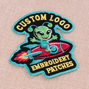 May include: Embroidered patch featuring a green alien riding a red rocket. The patch has the words "CUSTOM LOGO" and "EMBROIDERY PATCHES" in yellow lettering on a black background with a teal border. The alien is smiling.