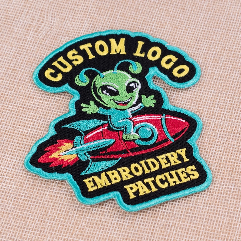 May include: Embroidered patch featuring a green alien riding a red rocket. The patch has the words "CUSTOM LOGO" and "EMBROIDERY PATCHES" in yellow lettering on a black background with a teal border. The alien is smiling.