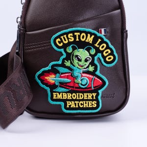May include: A brown leather crossbody bag with a colorful embroidered patch. The patch features a green alien on a red rocket ship with the text "CUSTOM LOGO" and "EMBROIDERY PATCHES". The bag has a shoulder strap.