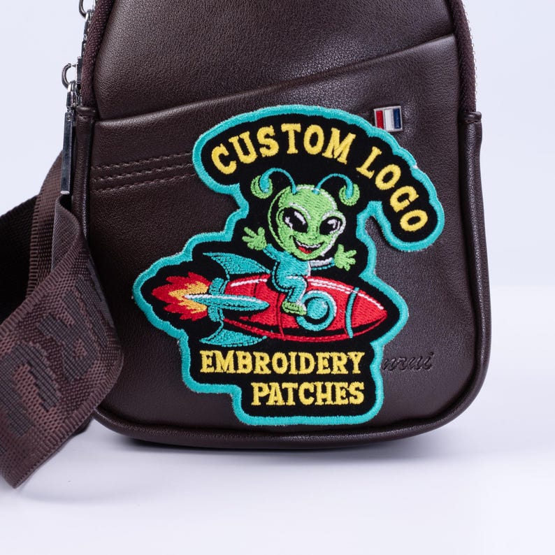 May include: A brown leather crossbody bag with a colorful embroidered patch. The patch features a green alien on a red rocket ship with the text "CUSTOM LOGO" and "EMBROIDERY PATCHES". The bag has a shoulder strap.