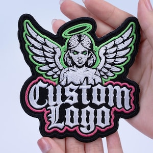 May include: Embroidered patch featuring an angel with green eyes and a halo. The angel has white wings with green trim. The text "CUSTOM LOGO" is in white gothic lettering, outlined in pink, on a black background.