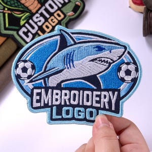 May include: An oval-shaped embroidered patch featuring a light blue shark graphic centered within a dark blue border. Two black and white soccer balls are positioned on the left and right sides. The text EMBROIDERY LOGO is stitched in white.